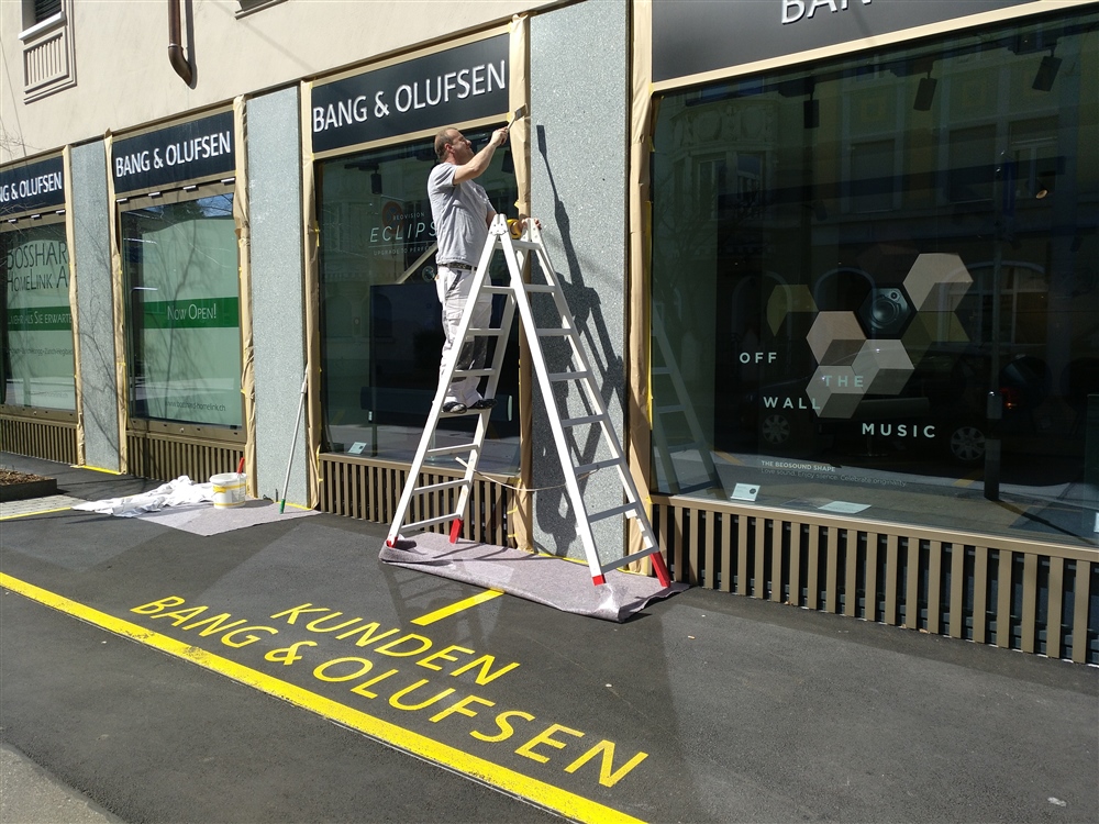 Maler beschriftet ein Schaufenster, davor eine gelbe Bodenmarkierung